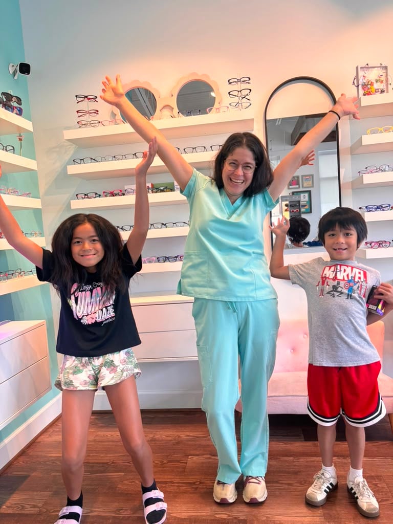 Dr. Teten and two smiling kids raise their arms in celebration in front of eyewear displays at Navigation Eye Care.
