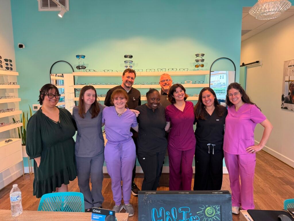 The Navigation Eye Care team smiles together in the practice's teal showroom, with eyewear on display behind them.