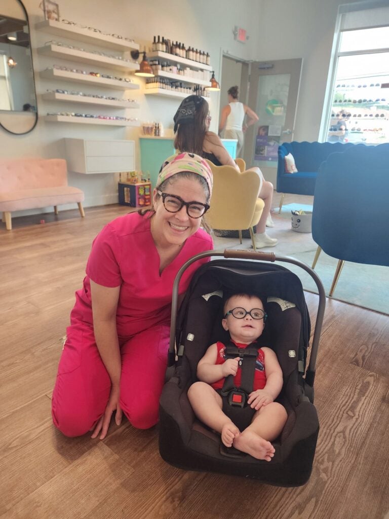 Dr. Teten, an optometrist at Navigation Eye Care, smiles while kneeling beside an infant patient seated in a car seat, both wearing glasses, inside the office with eyewear displays and colorful waiting area furniture in the background.